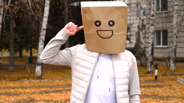 Uomo sorridente che indossa un sacchetto di carta con emozione felice, uomo in maschera in piedi all'aperto e dito vorticoso alla tempia — Video Stock