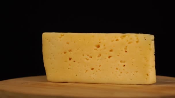 Fast rotation of piece of yellow cheese with holes on wooden board on black background. — Vídeo de Stock