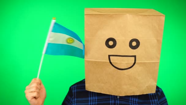Portret mężczyzny z papierową torbą na głowie machającego argentyńską flagą z uśmiechniętą twarzą na zielonym tle. — Wideo stockowe