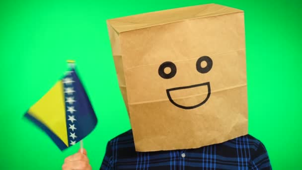 Portrait of man with paper bag on head waving Bosnia and Herzegovina flag with smiling face against green background. — Stock Video