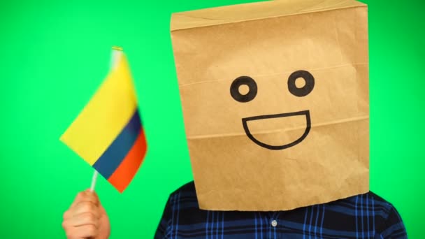 頭の上に紙の袋を持つ男の肖像緑の背景に笑顔でコロンビアの旗を振って. — ストック動画