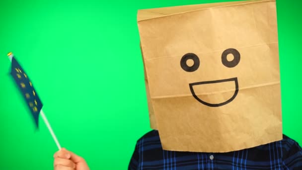 Portret van de mens met papieren zak op hoofd zwaaiend met Europese vlag met lachend gezicht tegen groene achtergrond. — Stockvideo