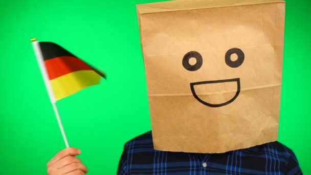 Portret van een man met papieren zak op hoofd zwaaiend met Duitse vlag met lachend gezicht tegen groene achtergrond. — Stockvideo