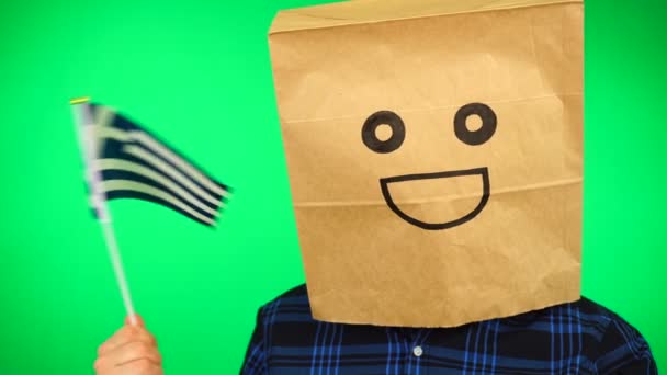 Portret van de mens met papieren zak op hoofd zwaaiend met Griekse vlag met lachend gezicht tegen groene achtergrond. — Stockvideo