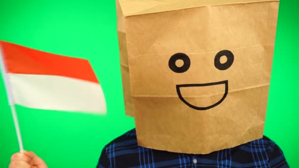 頭の上に紙の袋を持つ男の肖像緑の背景に笑顔でインドネシアの旗を振って. — ストック動画