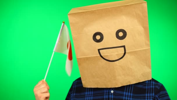 Ritratto di uomo con sacchetto di carta sulla testa sventola bandiera giapponese con volto sorridente sullo sfondo verde. — Video Stock