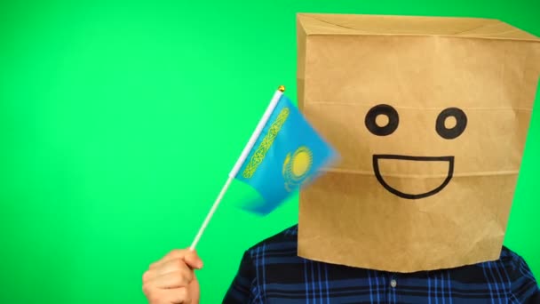 Portrait of man with paper bag on head waving Kazakhstan flag with smiling face against green background. — Stock Video