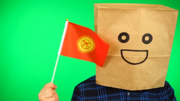 Portrait d'homme avec sac en papier sur la tête agitant drapeau kirghize avec visage souriant sur fond vert. — Video