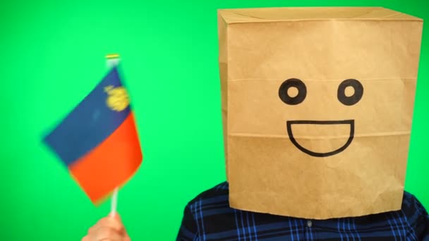 Portrait of man with paper bag on head waving Liechtenstein flag with smiling face against green background. — Stock Video