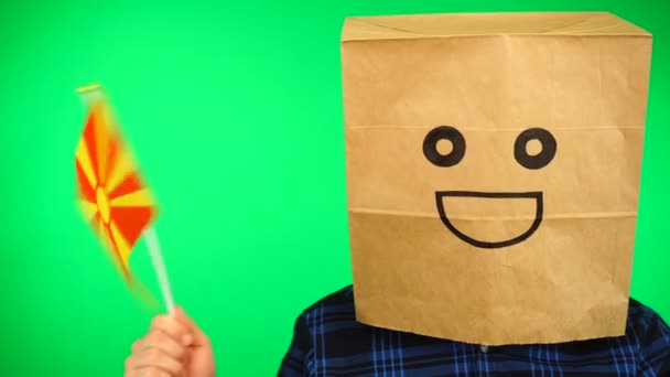 Portrait of man with paper bag on head waving Macedonian flag with smiling face against green background. — Stock Video