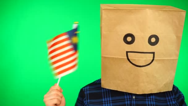Porträtt av man med papperspåse på huvudet viftande malaysiska flagga med leende ansikte mot grön bakgrund. — Stockvideo