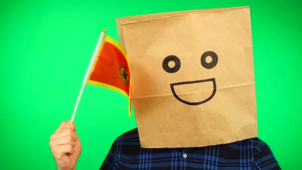 Retrato de hombre con bolsa de papel en la cabeza ondeando bandera montenegrina con la cara sonriente sobre fondo verde. — Vídeo de stock