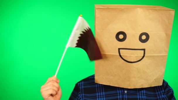 Portrait of man with paper bag on head waving Qatari flag with smiling face against green background. — Stock Video