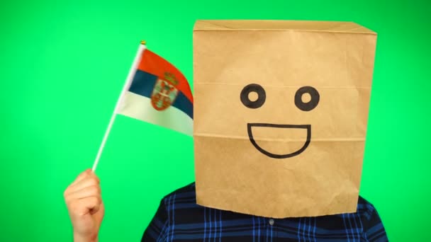 Portrait of man with paper bag on head waving Serbian flag with smiling face against green background. — Stock Video