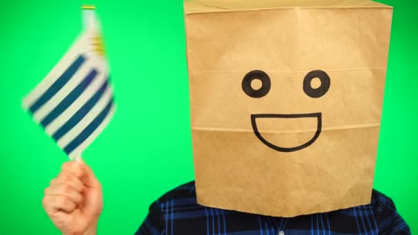 Kafasında kağıt torba olan ve yeşil arka planda gülümseyen Uruguay bayrağı sallayan bir adamın portresi.. — Stok video