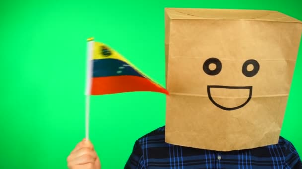 Portret van man met papieren zak op hoofd zwaaiend Venezolaanse vlag met lachend gezicht tegen groene achtergrond. — Stockvideo