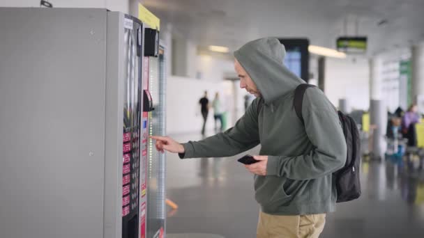 Un uomo con uno zaino all'aeroporto. Il ragazzo paga con il suo smartphone per il suo acquisto nel distributore automatico. — Video Stock
