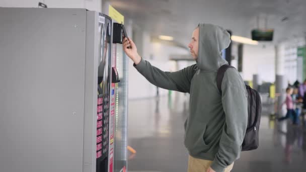 Un hombre con una mochila en el aeropuerto. El tipo paga con su smartphone por su compra en la máquina expendedora. — Vídeo de stock