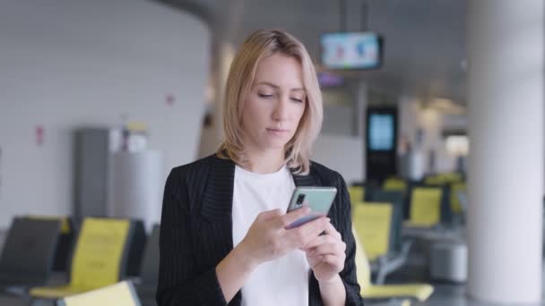 En attraktiv ung kvinna på flygplatsen sms:ar med vänner på sin smartphone. — Stockvideo