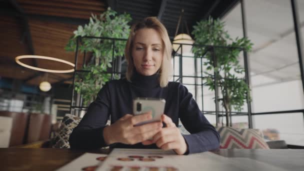 Mulher loira atraente usa um smartphone em um café. Uma mulher está conversando com amigos e colegas. — Vídeo de Stock