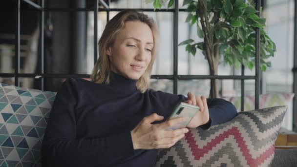 Uma loira elegante em um restaurante usa um smartphone. Uma mulher está conversando com amigos nas redes sociais. — Vídeo de Stock