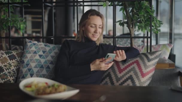 Μια κομψή ξανθιά σε ένα εστιατόριο χρησιμοποιεί ένα smartphone. Μια γυναίκα συνομιλεί με φίλους στα κοινωνικά δίκτυα.. — Αρχείο Βίντεο