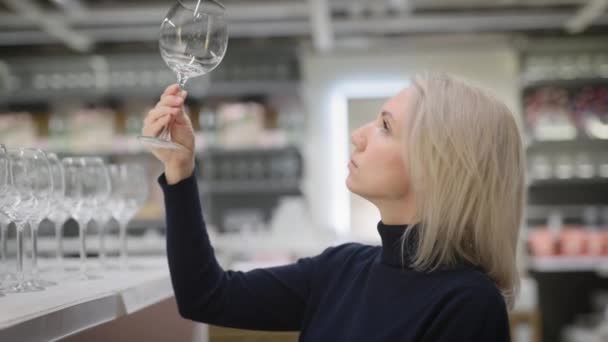 Elegant blondin i en köksbutik. En kvinna väljer nya vinglas till en fest. — Stockvideo
