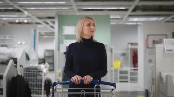 Una mujer en una gran tienda de mejoras para el hogar. Una mujer rubia con un carrito de compras camina a través de una gran sala de exposición. — Vídeo de stock