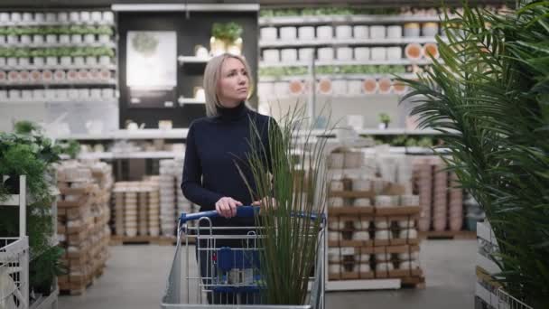 Mooie jonge vrouw in een plantenwinkel. De blonde kiest voor landschapsarchitectuur voor het huis. — Stockvideo
