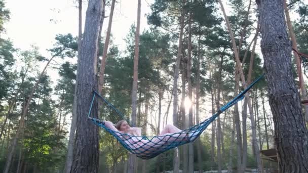 Het meisje ligt in een hangmat, rustend in een dennenbos. Ochtend eerste zonnestralen. — Stockvideo