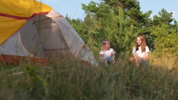 Dos chicas están sentadas cerca de la tienda y hablando. Campamento de verano en el bosque. — Vídeos de Stock