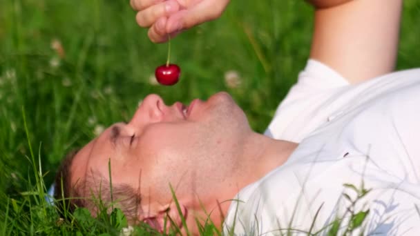 Homem relaxante deitado na grama no jardim e comer suculentas cerejas maduras — Vídeo de Stock