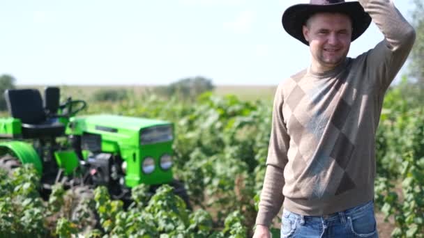 トラクターの後ろに立った帽子を被った若い農民がカメラに微笑んだ。. — ストック動画