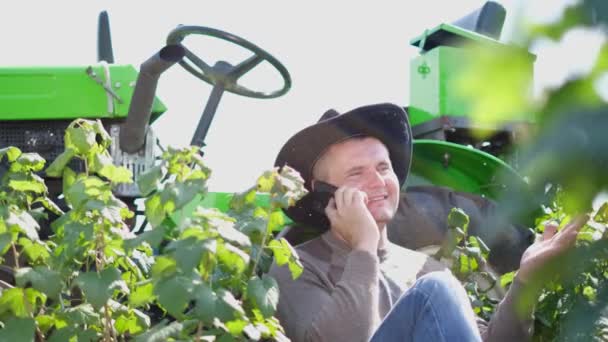 Un joven agricultor usa un teléfono inteligente, está descansando cerca de su tractor. — Vídeos de Stock