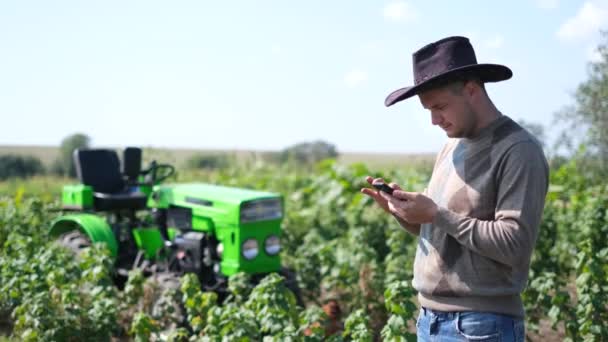 Ein junger Landarbeiter hält ein Smartphone in der Hand, während er in der Nähe seines Traktors arbeitet. — Stockvideo