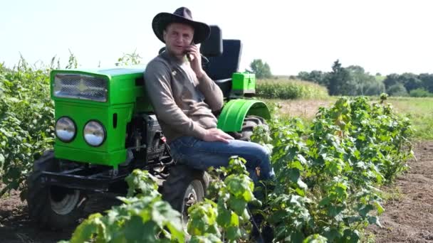 Un joven agricultor usa un teléfono inteligente, está descansando cerca de su tractor. — Vídeos de Stock
