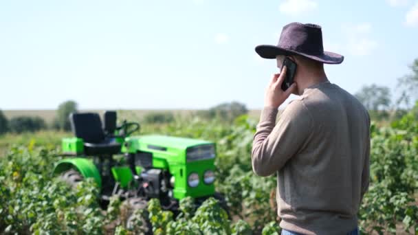 En ung bonde använder en smartphone, han vilar nära sin traktor. — Stockvideo