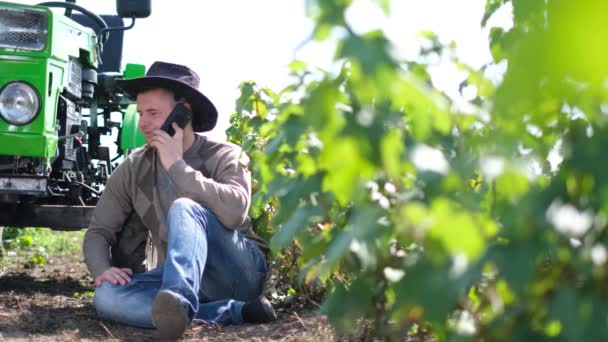 A young agricultural worker holds a smartphone while he is working near his tractor. — Stock Video