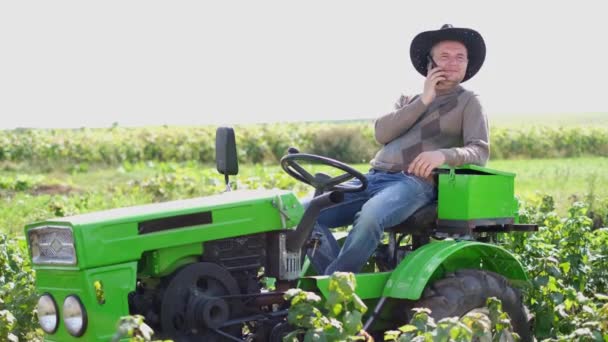 Un joven granjero descansando sentado en un tractor verde. Hombre del pueblo hablando por teléfono. — Vídeo de stock