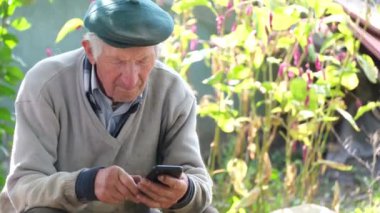 Beyaz saçlı büyükbaba evin yanındaki sandalyede otururken akıllı telefon kullanıyor..