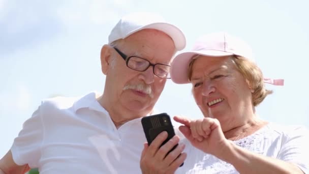 Avós modernos usam um smartphone, eles se divertem sentados em um parque da cidade — Vídeo de Stock