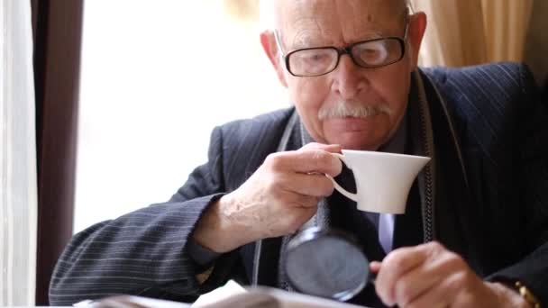 Starší muž drží šálek kávy sedí u stolu v kavárně a čte noviny. — Stock video