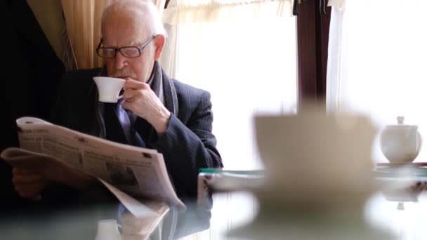 Un uomo anziano si siede su una sedia e legge un giornale davanti al caffè del mattino in un caffè. — Video Stock