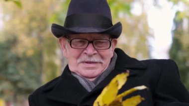 Gözlüklü ve siyah ceketli yaşlı ve yalnız büyükbaba, bir sonbahar parkında bankta oturuyor.