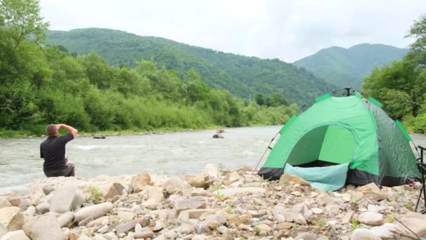 Un hombre de vacaciones junto a un río de montaña, armó una carpa verde. — Vídeo de stock