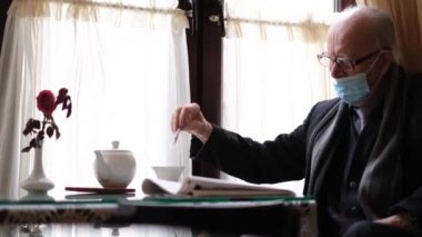 Koruyucu maskeli yaşlı bir emekli kafede otururken çayı karıştırıyor.