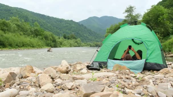 Turista postavil zelený stan poblíž horské řeky a odpočívá ve stanu.. — Stock video