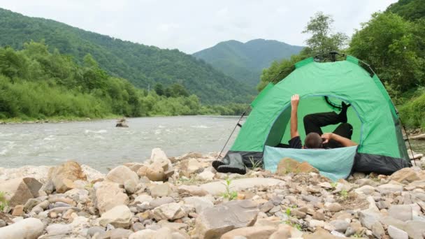 El turista puso una tienda verde cerca de un río de montaña y está descansando acostado en una tienda de campaña. — Vídeo de stock