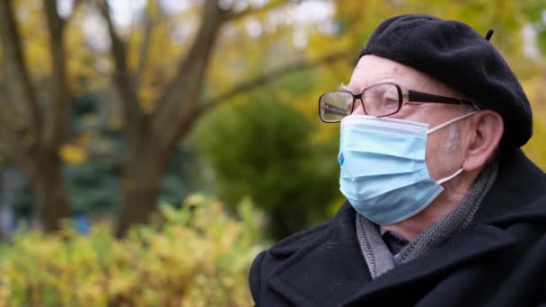 Um homem idoso com uma máscara protetora médica e óculos de proteção está descansando no parque. — Vídeo de Stock