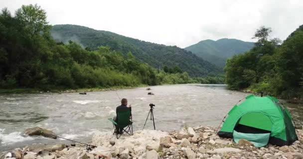 Um homem está pescando em um rio de montanha, ele está sentado em uma cadeira perto da tenda — Vídeo de Stock